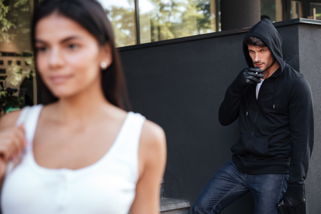 a white man wearing a hoodie and gloves, smoking a cigarette, looks at a young girl with long dark hair while she is unaware