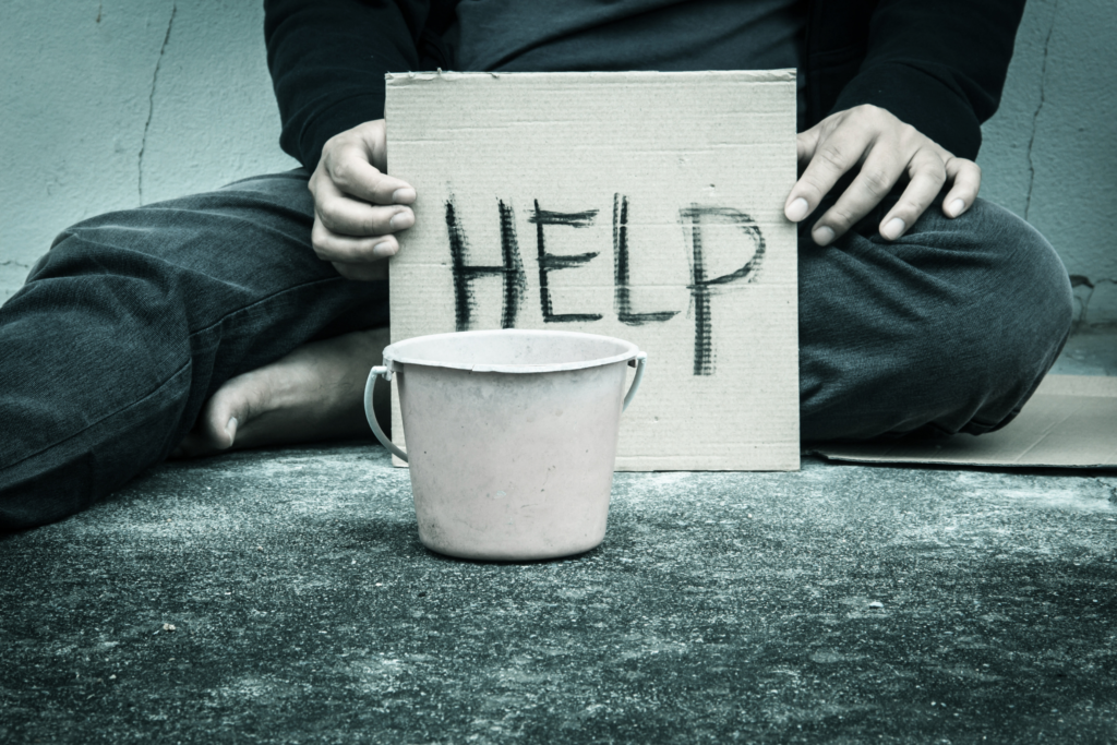 a white male sitting on concrete while holding a cardboard sign with the word HELP handwritten on it behind a money collection pail